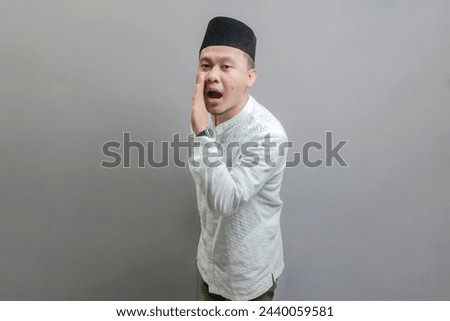 Happy asian muslim man hand on mouth telling secret rumor, whispering malicious talk conversation wearing a koko shirt and peci with shades of the fasting month, isolated on a gray background