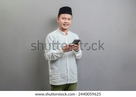 Happy asian muslim man using smart phone cellphone for calls, social media, mobile application online, wearing a koko shirt and peci with shades of the fasting month, isolated on a gray background