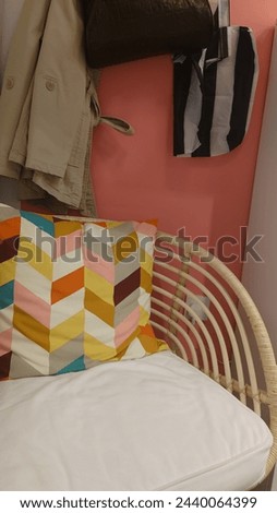 A colorful chevron cushion in the corner of an open, wicker chair with white cushions and hanging on it is a black handbag, while above hangs two jackets and below lies another folded coat. 