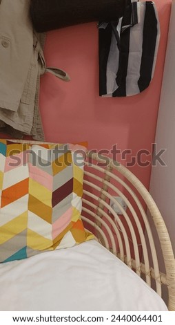 A colorful chevron cushion in the corner of an open, wicker chair with white cushions and hanging on it is a black handbag, while above hangs two jackets and below lies another folded coat. 