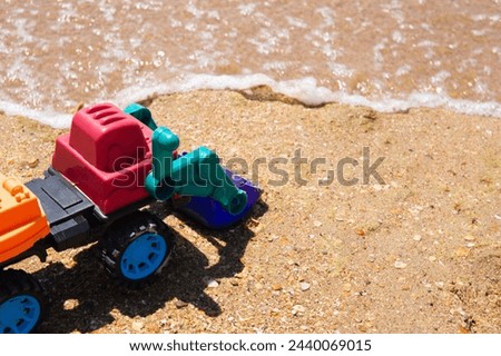 A toy excavator sits on sand, waves in the background. It's colorful, playful, family vacation, development crisis, safety rules near sea