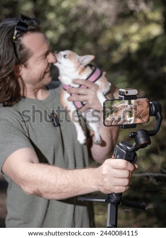 Man using a phone gimbal to film himself and a chihuahua he is holding. The film is happening in a natural forest location.