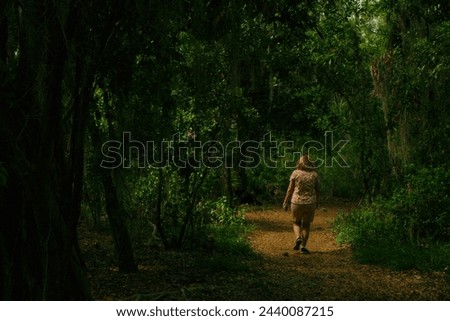 Older white lady on Leading line s curve view down a light brown dirt nature trail path layered in leaves with trees left and right making a canopy over the trail. Sunshine and shade on a sunny day.  Royalty-Free Stock Photo #2440087215