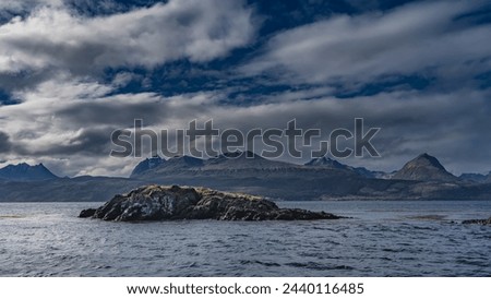 A small rocky island with sparse grassy vegetation is visible in the ocean. Picturesque coastal mountains of the Andes against a blue sky and clouds. Argentina. Tierra del Fuego Archipelago.
