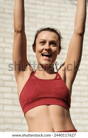 Caucasian fitness model showing happiness with her arms raised. She is in front of a white brick wall and she is wearing a red sport bra. Achieving goals and celebrate concept. Royalty-Free Stock Photo #2440117627