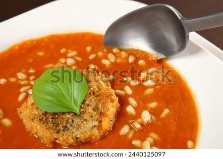 A picture of a white bowl full of fresh vegetable cream soup with pine nuts and piece of toast with basil on top