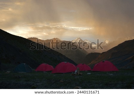 Picture of red colored camping tents in a green meadow surrounded by huge snow covered mountains and sunlight creeping in through the clouds. Sunrays showing Tyndall effect.