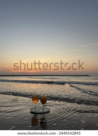 Waves rolling towards the beach, submerging the two fresh juice glass placed on a black trey which is lying by the beach during the sunset. The picture has a negative space.