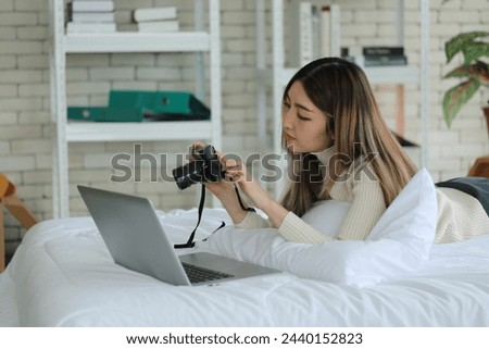 Asian woman lying on bed, happy working at home and holding camera, checking photos, contributors selling, stock photos on website, working online in apartment