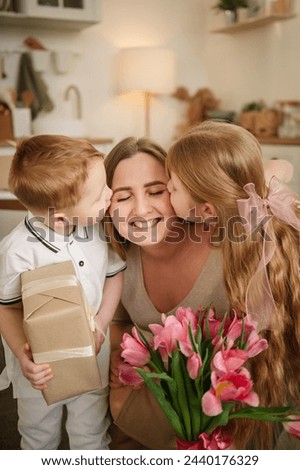 Little son and daughter hugging and kissing mom and make surprise for Mothers Day. Children give bouquet of tulip flowers. Childs gives present box congratulates surprised mom in kitchen. Happy family