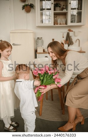 Little boy son holds and gives bouquet of tulip flowers. Daughter gives present box congratulates surprised mom in kitchen. Children make surprise for Mothers Day. Side view. Happy family holiday