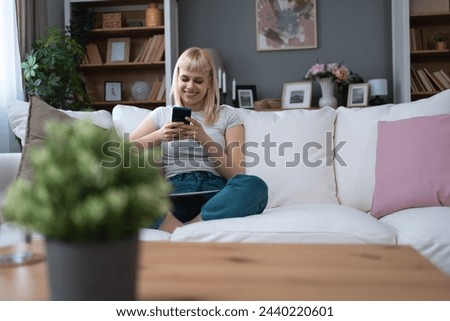 Happy millennial blonde teen girl checking social media holding smartphone at home Smiling young woman using mobile phone app playing game shopping online ordering delivery relax on sofa Online dating