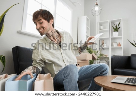 Young tattooed man with gift voucher and shopping bags at home