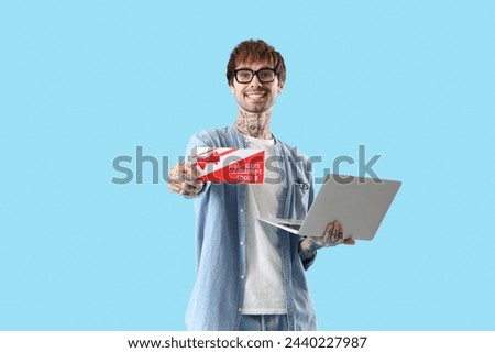 Young tattooed man with gift voucher and laptop on blue background