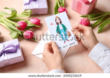 Female hands with kid drawing, gift boxes and tulips on beige grunge background. Happy Mother's Day