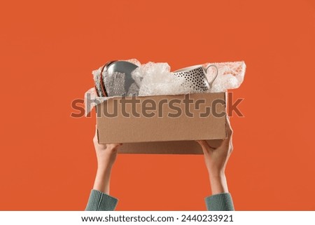 Woman with kitchen utensils in moving box on orange background
