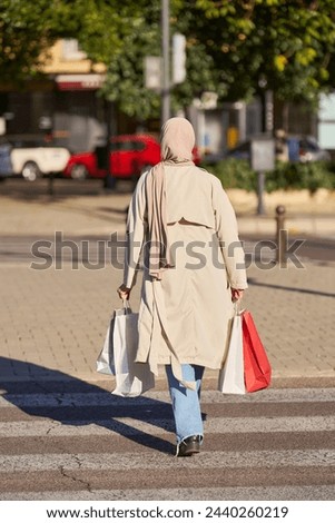 A woman wearing a hijab is walking across a crosswalk with shopping bags. The bags are red and white