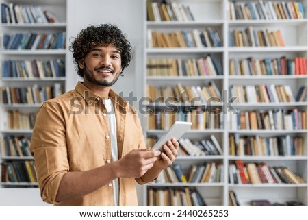 A cheerful young adult male holds a tablet and smiles confidently in a library with bookshelves in the background.