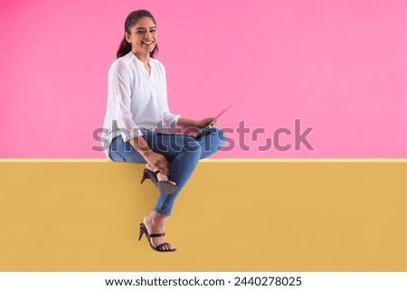 Woman with laptop on lap sitting on wall and looking at camera with smile Royalty-Free Stock Photo #2440278025