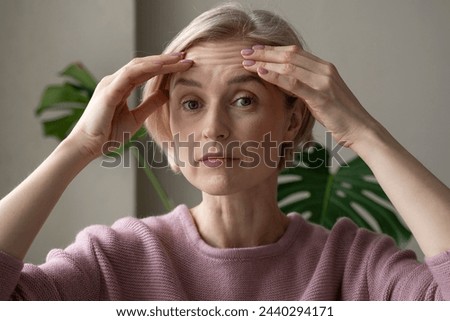 A contemplative mature woman is captured indoors while gently touching her forehead with both hands, appearing introspective or concerned. She wears a soft pink sweater, and her expression conveys Royalty-Free Stock Photo #2440294171