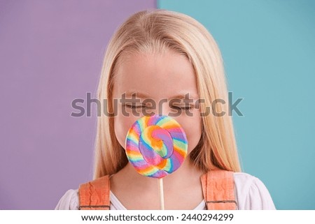 Child, lollipop and candy dessert in studio fr party snack at carnival or rainbow swirl, eating or sweets. Female person, happy and hiding as split purple or blue background for hungry, treats or fun Royalty-Free Stock Photo #2440294299