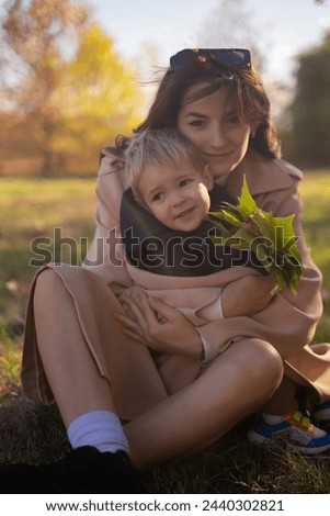 A mother and son embrace and play in the spring park. The boy holds green tree leaves while the mother tickles him, creating precious family moments on Mother's Day, enjoying a picnic in nature.