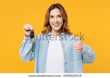 Young smiling happy woman she wear blue shirt white t-shirt casual clothes hold in hand car keys fob keyless system show thumb up isolated on plain yellow background studio portrait. Lifestyle concept