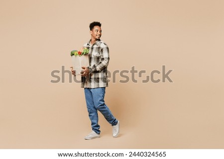 Full body sideways profile young man wear grey shirt hold paper bag for takeaway mock up with food products go look aside isolated on plain beige background. Delivery service from shop or restaurant
