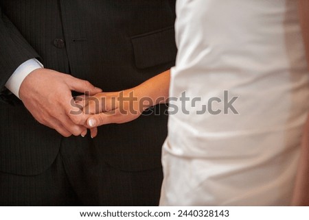 This close-up captures the tender moment where a bride places a wedding ring on the groom's finger. Their hands meet against the contrasting fabrics of the groom's suit and the bride's dress, a