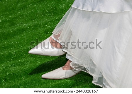 A bride's classic pointed white wedding shoes peek from beneath the soft layers of a flowing gown against a lush green artificial turf, blending traditional bridal style with a touch of modernity