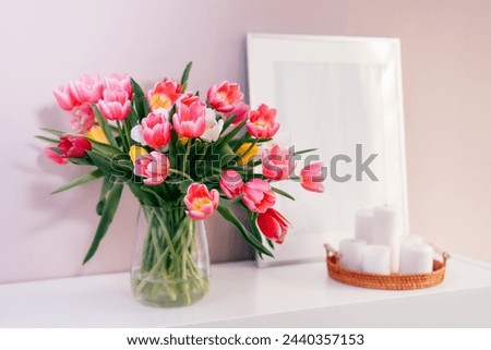 Modern interior design with focus on tulip flowers bouquet in vase, white picture frame mockup, candles on white console on pink wall background. Poster design mockup. Minimalist Home decor.