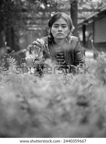 Intense mixed race woman looking at camera in a monochrome portrait. There are blurry vegetation in the foreground and a blurry building in the background. Medium format analog film camera.