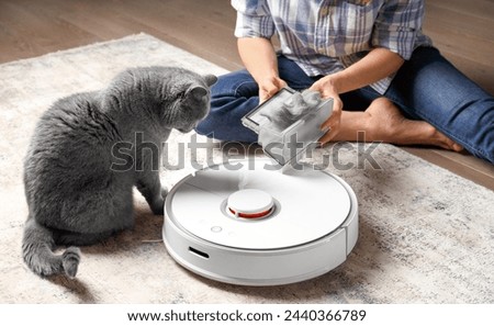 The container of the vacuum cleaner robot is in female hands, the dust collector of the robot vacuum cleaner is full of wool, the cat sits nearby and looks at the container. Royalty-Free Stock Photo #2440366789