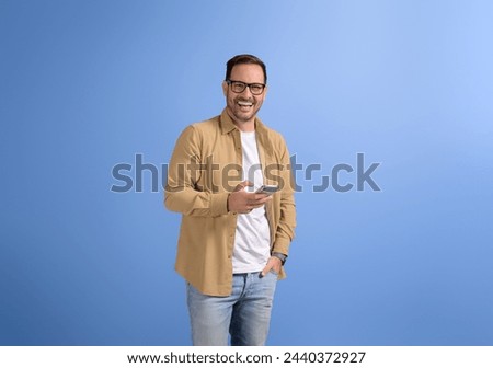 Young happy businessman with hand in pocket using social media on smart phone over blue background