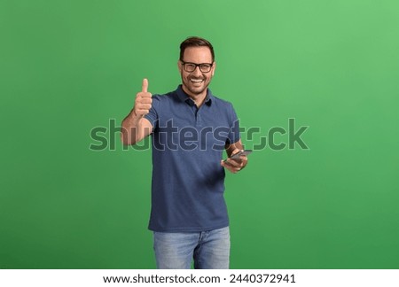 Positive young businessman with smart phone showing thumbs up sign while posing on green background