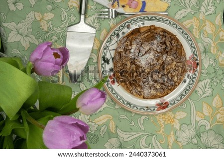 Pear cake with walnuts and powdered sugar on a plate. Fresh green patterned tablecloth, cake fork and cake server. Purple tulips protrude into the picture. Shot from above