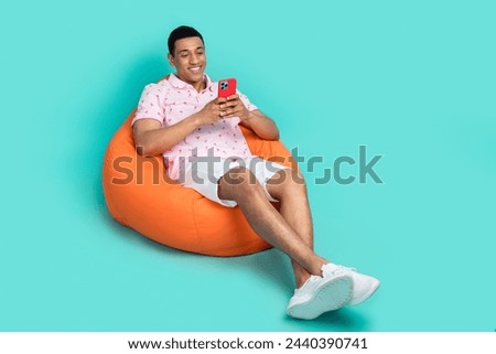 Full size photo of optimistic guy dressed print t-shirt sit on pouf look at smartphone read email isolated on teal color background