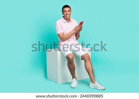Full size photo of attractive smart guy dressed polo white shorts sit on podium holding smartphone isolated on teal color background