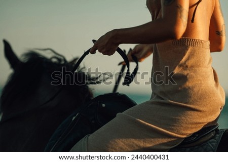 Close up of a tattooed woman holding a lead while riding a horse in wild at summertime at sunset. Cropped picture of female's hands holding a leash and horseback riding near water in nature. Close up.