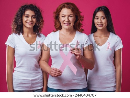 Women against breast cancer. Beautiful women of different ages and nationalities with pink ribbons on their chests smiling, one is holding a pink paper ribbon, on red background