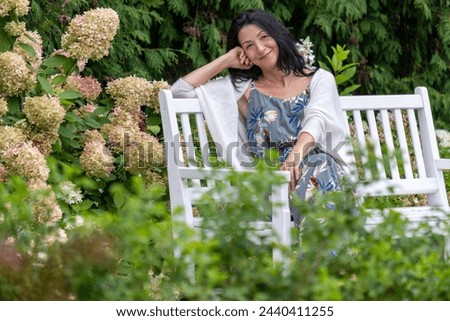 In a lush garden, a woman with a contented smile rests on a bench, enveloped in greenery and the soft hues of hydrangeas, a picture of serene leisure. High quality photo