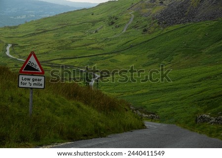 Traffic sign announces a steep descent down a winding road through green hills. A warning sign for a steep road ahead on a serpentine mountain road in the middle of a hilly landscape in Lake District.
