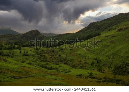 AERIAL: Threatening rain clouds hover above rolling green hills of Lake District, hinting at an upcoming downpour in a peaceful natural environment. Moody weather in a picturesque mountain region.