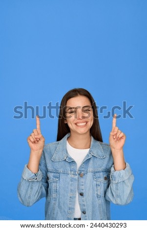 Happy cute Latin teenage student girl pointing up standing on background. Smiling pretty hispanic teenager with brunette hair wearing denim jacket presenting ads standing isolated on blue. Vertical Royalty-Free Stock Photo #2440430293