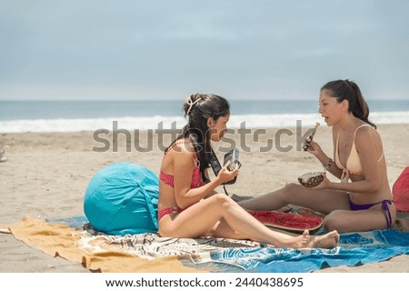 Two latin young women wearing bikini sitting under umbrella enjoying picnic on the beach
