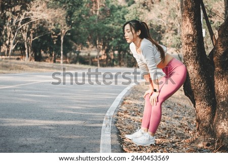 A runner takes a well-deserved break, leaning against a tree as she catches her breath on a scenic park trail, highlighting a moment of rest in her fitness journey. Royalty-Free Stock Photo #2440453569