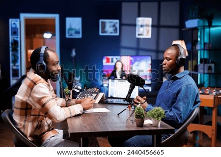 Sitting at table using the audio equipment are two african american men discussing and addressing their online audience. Interviewer asking male influencer questions, recording a radio show at home.