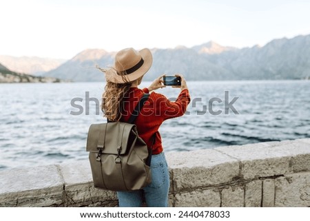 Female traveler in casual sweater taking picture of amazing landscape with mountains, ocean and sunset sky on smartphone during vacation.Tourist during holidays.