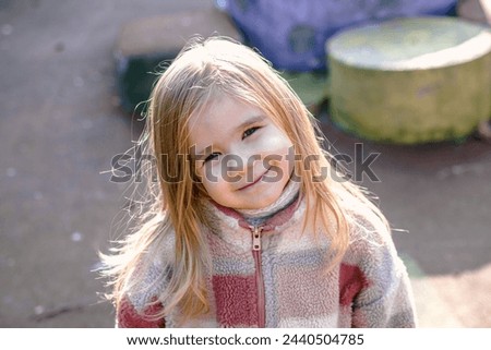 Smiling young girl wearing a stylish checkered jacket, posing for the camera with a blurred outdoor background of trees. She exudes joy and happiness in the natural setting.