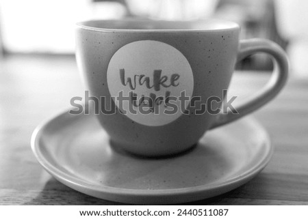 Wake up, message of the brand new day. Sign letters. Close up detail of coffee cup. Blurry background. Black and white, monochrome.